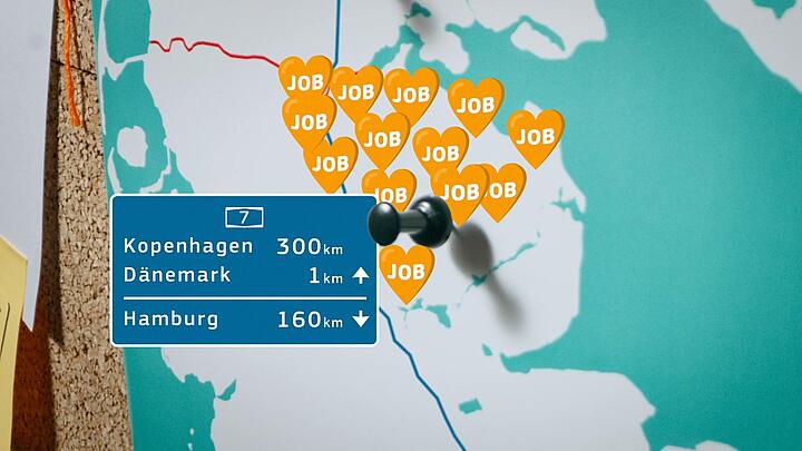 Eine Karte des Kreisgebiets Flensburg/Schleswig, auf dem Herzchen mit dem Wort "Job" verteilt sind und eine Grafik die Entfernungen zu Ballungszentren zeigt. Die Karte hängt an einer Pinnwand.